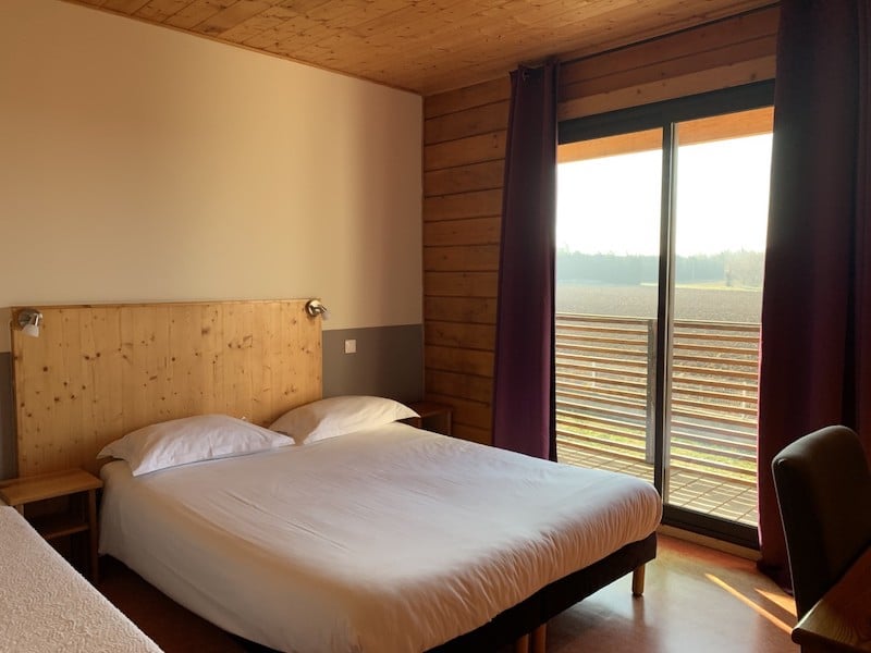Chambre Bois et Lumière avec Vue sur Campagne Chambre d'hôtel lumineuse avec un grand lit, tête de lit en bois clair, et une baie vitrée donnant sur un paysage de champs et un balcon à lattes horizontales.