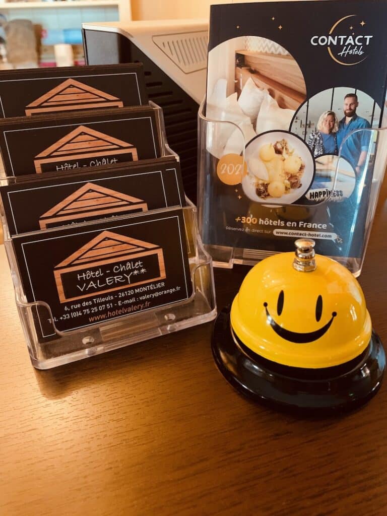 Hôtel Valéry : Cartes, Contact Hotels et sonnette Comptoir d'hôtel avec porte-cartes Hôtel-Chalet Valéry, un dépliant Contact Hotels et une sonnette jaune souriante.