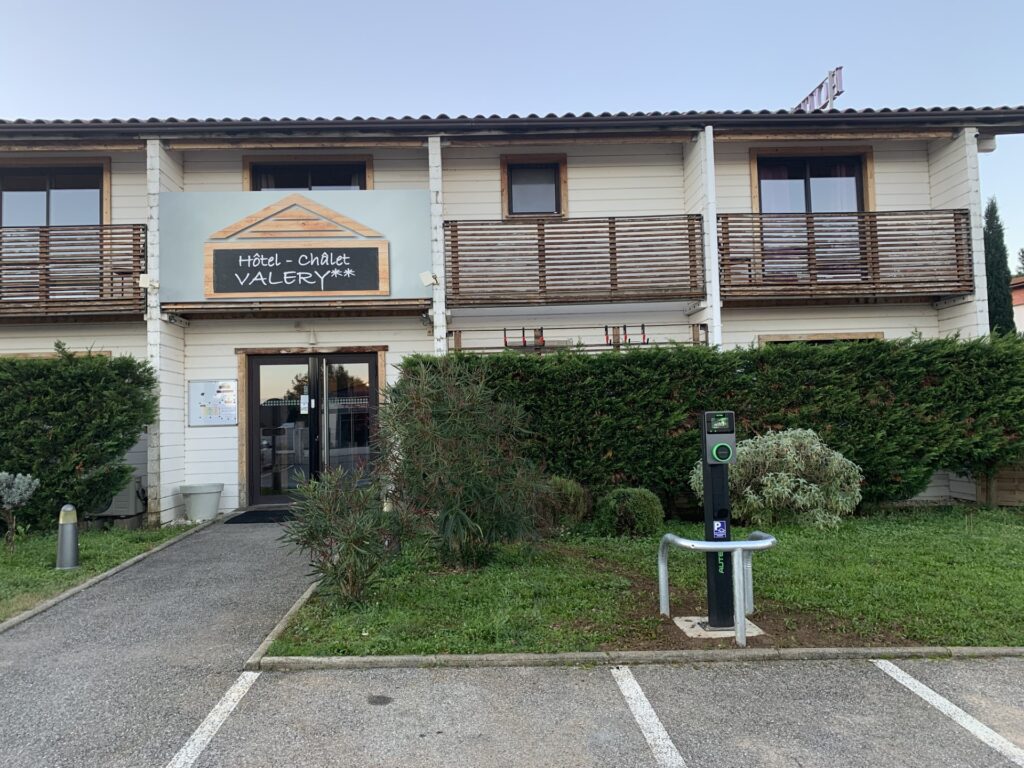 Hôtel Chalet Valery : borne de recharge et façade Façade de l'Hôtel - Chalet Valery** blanc avec balcons en bois, signalétique de borne de recharge EV et allée.