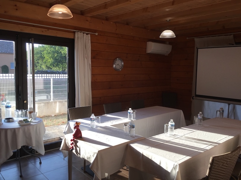 Salle de réunion boisée pour formation avec écran Salle de réunion ou de formation en bois, éclairée par la lumière du jour entrant par de larges portes vitrées, avec tables nappées et bouteilles d'eau.
