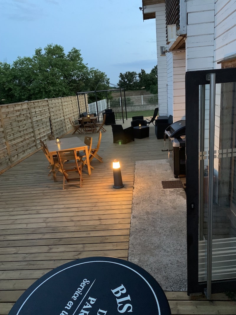 Terrasse de Soirée en Bois avec Mobilier et Lumières Terrasse en bois avec tables et chaises de jardin en soirée, éclairée par des lampes. Un panneau rond noir visible en bas porte l'inscription "BIS PAR... Service en te".