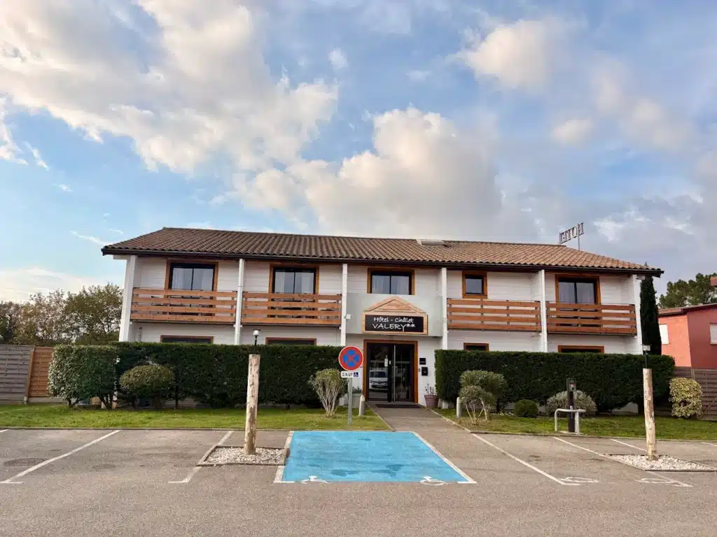 Hôtel Chalet Valery ** : Façade moderne et parking extérieur. Façade de l'Hôtel * Chalet VALERY**, bâtiment blanc à deux niveaux avec balcons en bois. Parking extérieur avec place PMR bleue, ciel nuageux.